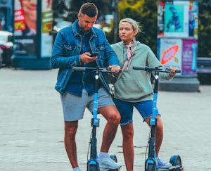 Scooter Deck: A man and woman are engaged with a phone while standing on their scooter decks, in the midst of an urban setting. The scooter deck is blue. Each scooter deck is flat.