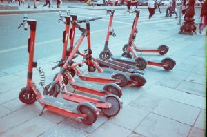 Scooter Deck: The image depicts a row of red electric scooters parked on a sidewalk, with their decks aligned and handles pointing upwards, ready for use. The color of each scooter deck is the same. Each scooter deck is flat.