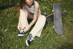 Skateboard: A person tying their shoes on the grass next to a skateboard, awaiting a skate session. She is sitting on another skateboard.