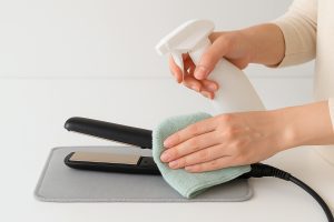 Hands cleaning flat iron plates with microfiber cloth and spray on a clean, minimal countertop.
