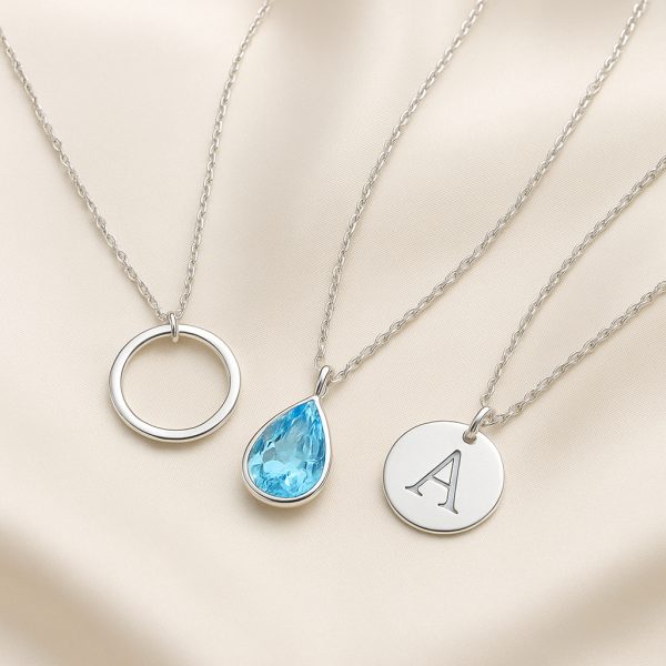 Sterling silver necklaces with open-circle, teardrop aquamarine, and initial disc pendants on ivory silk.