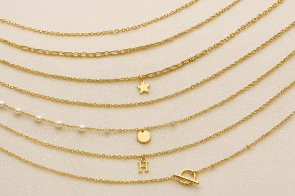 Assorted gold anklets in various chain styles with different clasps and small charms, elegantly arranged on ivory linen.