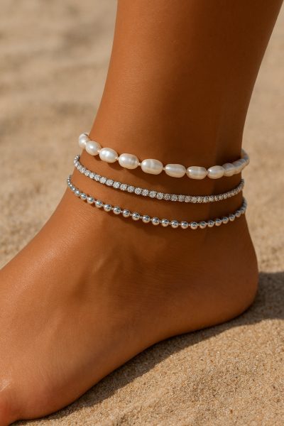 Stacked sterling silver anklets with pearls, zirconia, and beads on a model\u2019s ankle over sunlit sand.