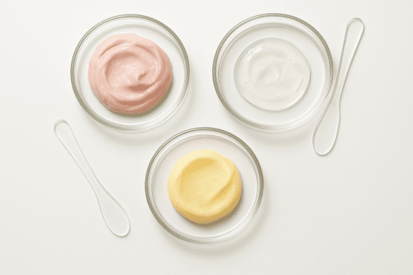 Overhead view of three glass dishes containing creamy, oily, and buttery lip mask formulas with spatulas on a white surface.