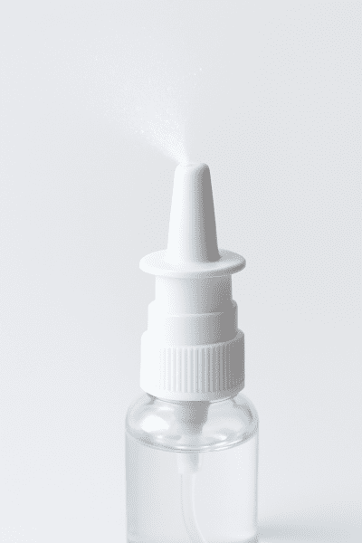 Macro image of a nasal spray bottle nozzle releasing a fine mist, showing intricate pump parts and a transparent solution in a sterile, white setting.